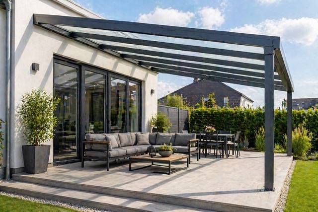 Patio area attached to a house with a glass-roof pergola, featuring a grey sectional sofa, coffee table, and a dining table with chairs on a tiled deck, surrounded by greenery.